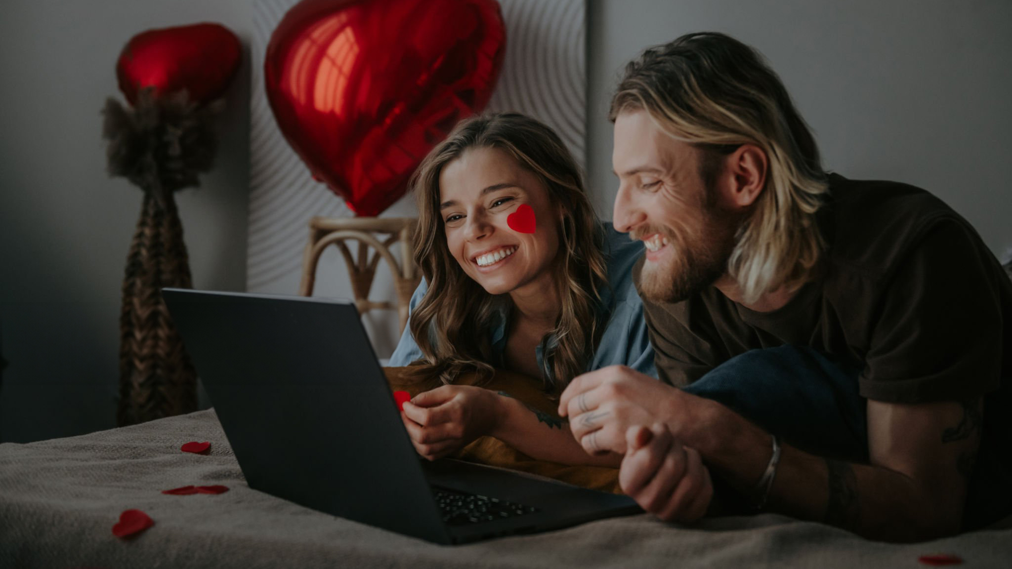 Couple playing a mystery romance game together on a laptop, with heart balloons and rose petals on the bed